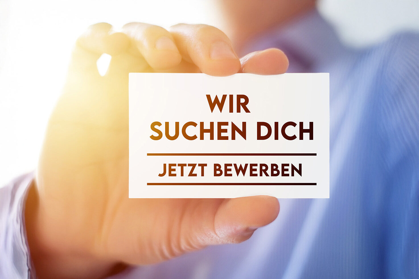 Ein Mann hält ein Schild mit der Aufschrift "Wir suchen Dich - Jetzt bewerben" in die Kamera.